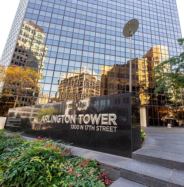 A glass skyscraper with several surrounding building reflected in the glass with a sign that says "Arlington Tower" in the foreground