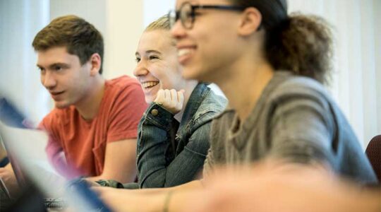 Three students smile while listening to a course lecture in a Khoury classroom