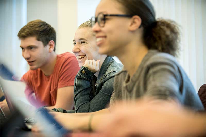 Three students smile while listening to a course lecture in a Khoury classroom