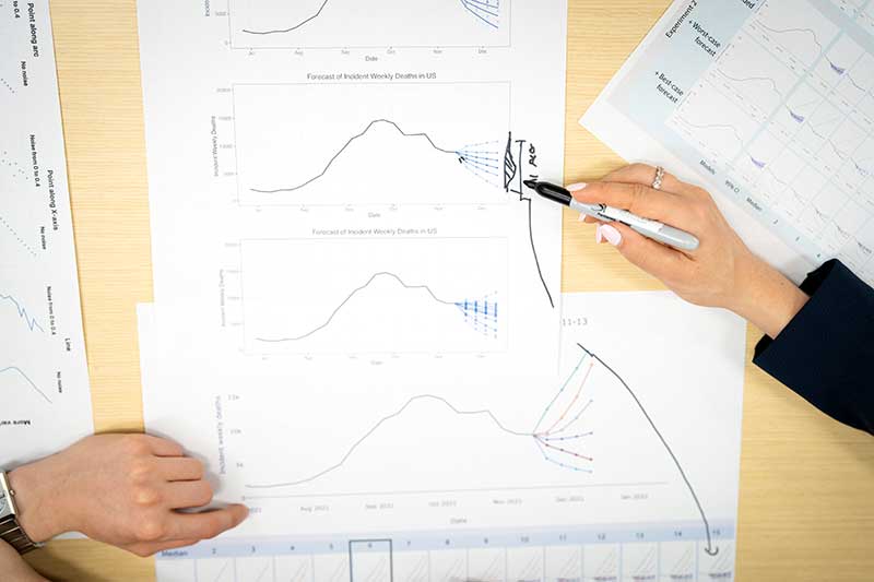 A table's surface is covered with several papers containing line charts. There are two hands pointing at different graphics.