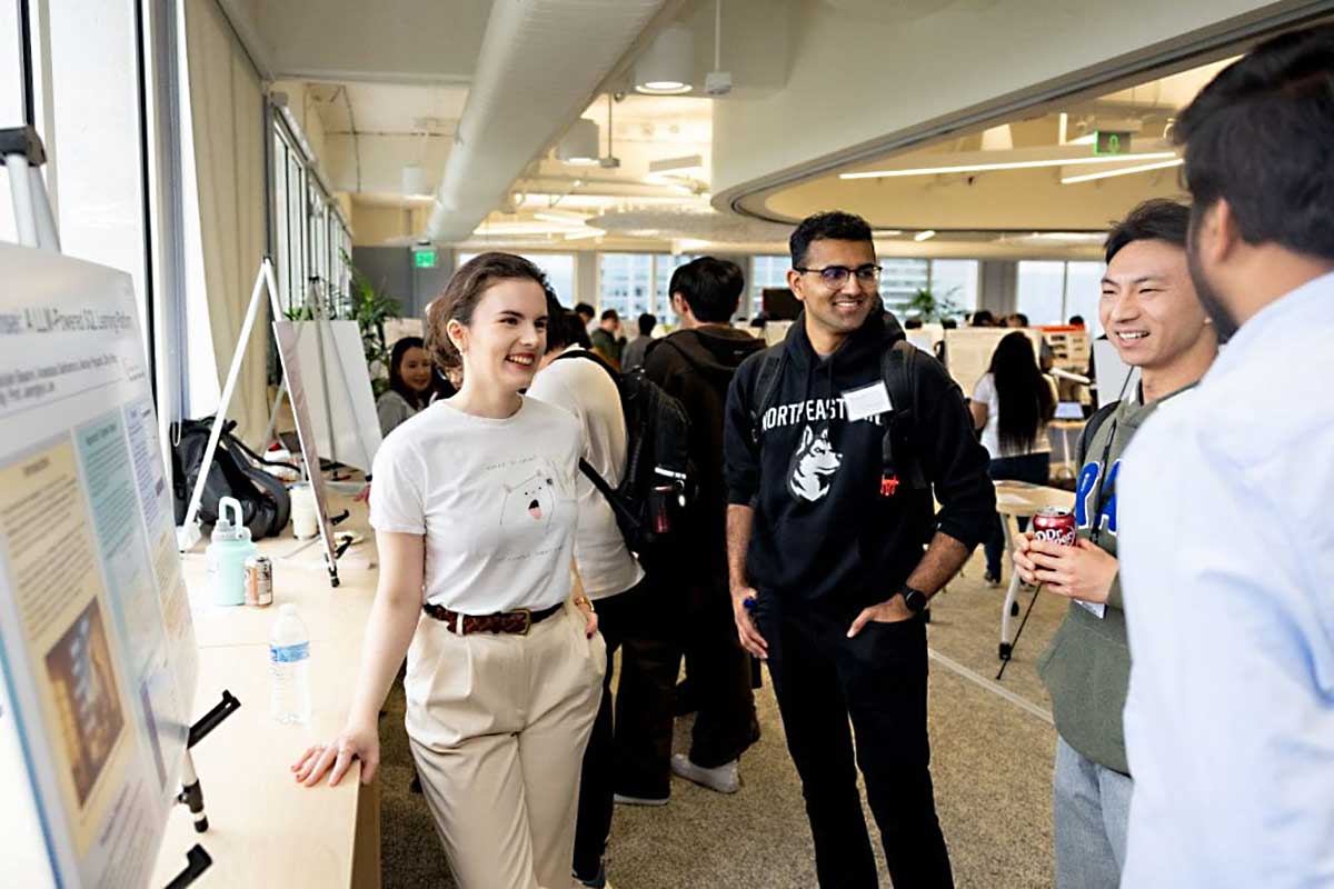 A student smiles standing next to her research poster while a fellow student asks her about the project