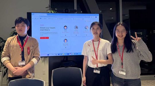 Three Khoury students stand in front of a large screen displaying team members who worked on a website to connect current Khoury students with Khoury alumni working in the students’ desired career field