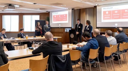 A team of students presents a project in a large conference room with judges viewing from a large u-shaped set of tables